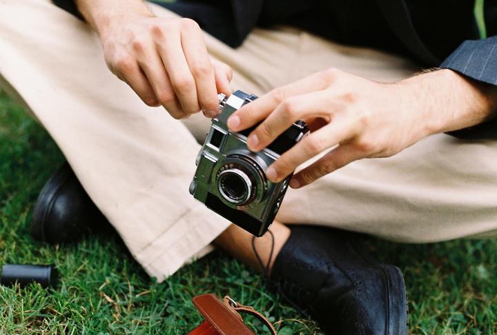 A man sits on the grass, holding a camera in his hands.