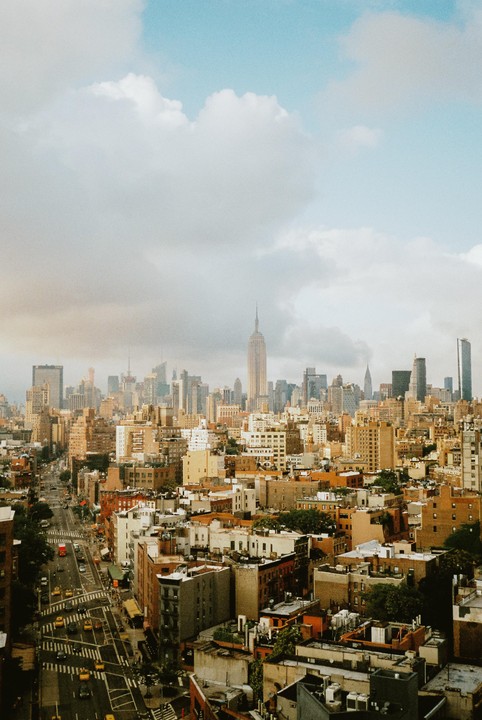 An aerial view shows a city street with cars and buildings, with the Empire State Building visible in the distance under a cloudy sky.
