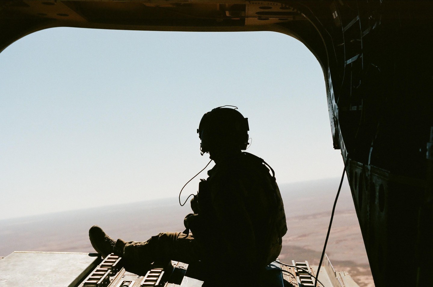 A person in military gear sits in the open doorway of an aircraft, looking out at a vast landscape below.