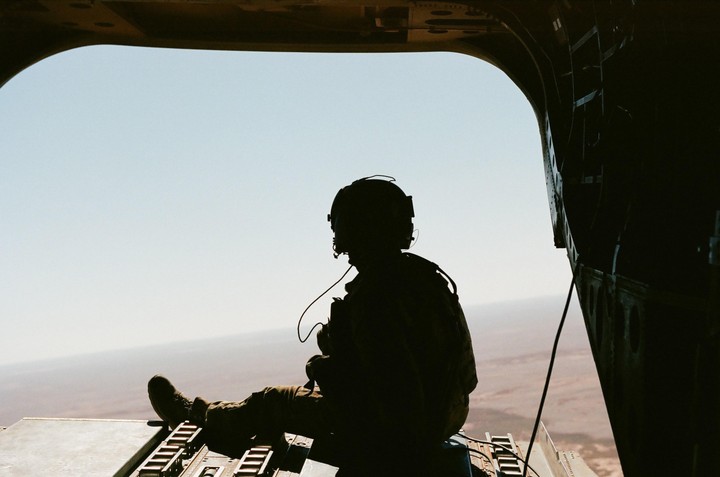 A person in military gear sits in the open doorway of an aircraft, looking out at a vast landscape below.