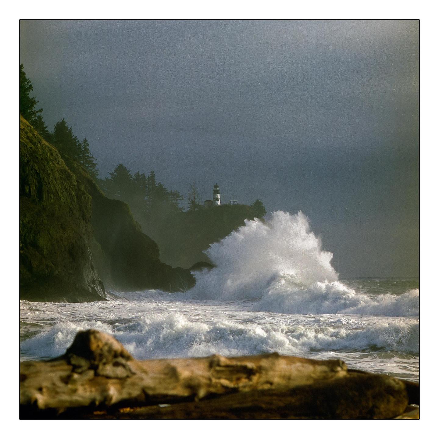 A large wave crashes against a rocky cliff with a lighthouse and trees on top under a dark sky.