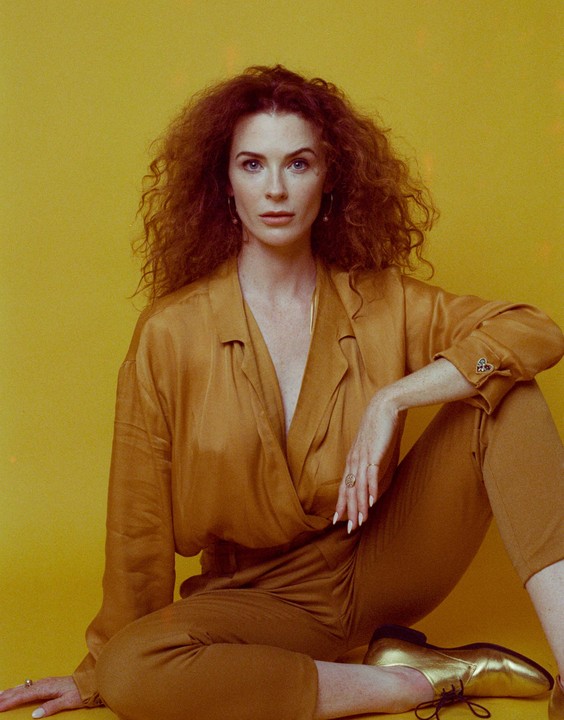 A woman with curly red hair and a gold jumpsuit sits against a yellow background.
