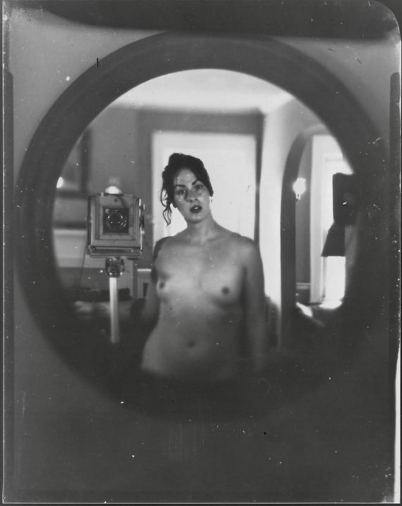A topless woman is reflected in a round mirror, with a large format camera visible to her left.