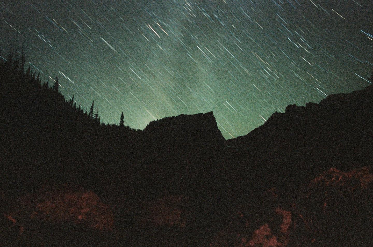 A long exposure photograph captures star trails above a dark mountain landscape with silhouetted trees.
