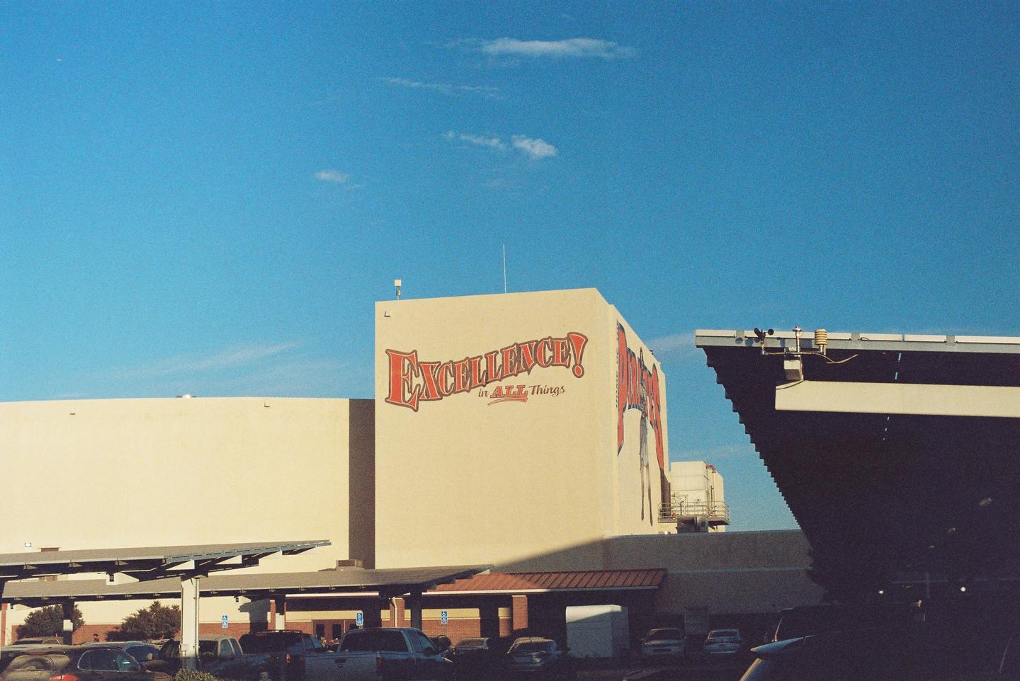 A building with "EXCELLENCE! in ALL Things" painted on its side stands next to a parking lot with solar panel awnings.