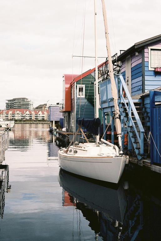 A white sailboat is docked next to colorful houses on stilts, with reflections visible in the water.