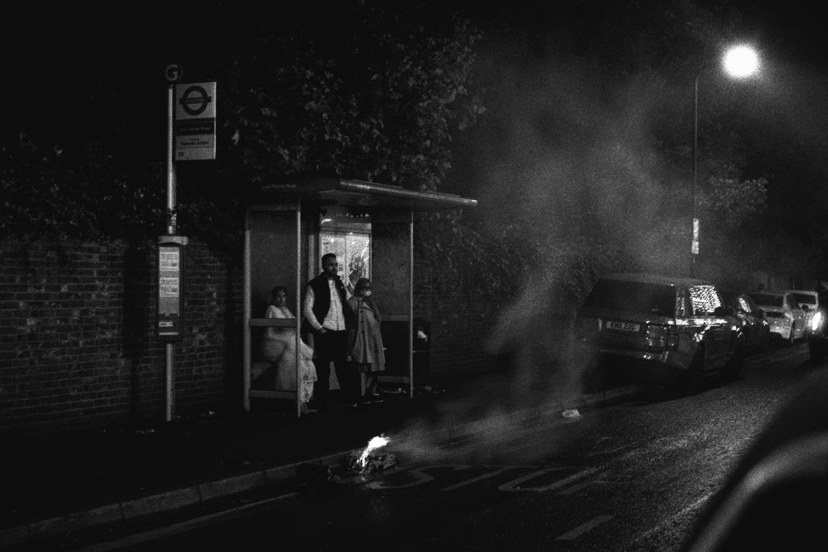 A man, woman, and child stand in a bus stop at night next to a small fire on the street.