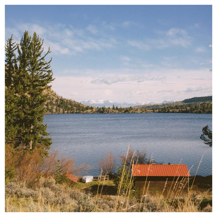 A small cabin with a red roof sits on the shore of a lake, with mountains in the distance.