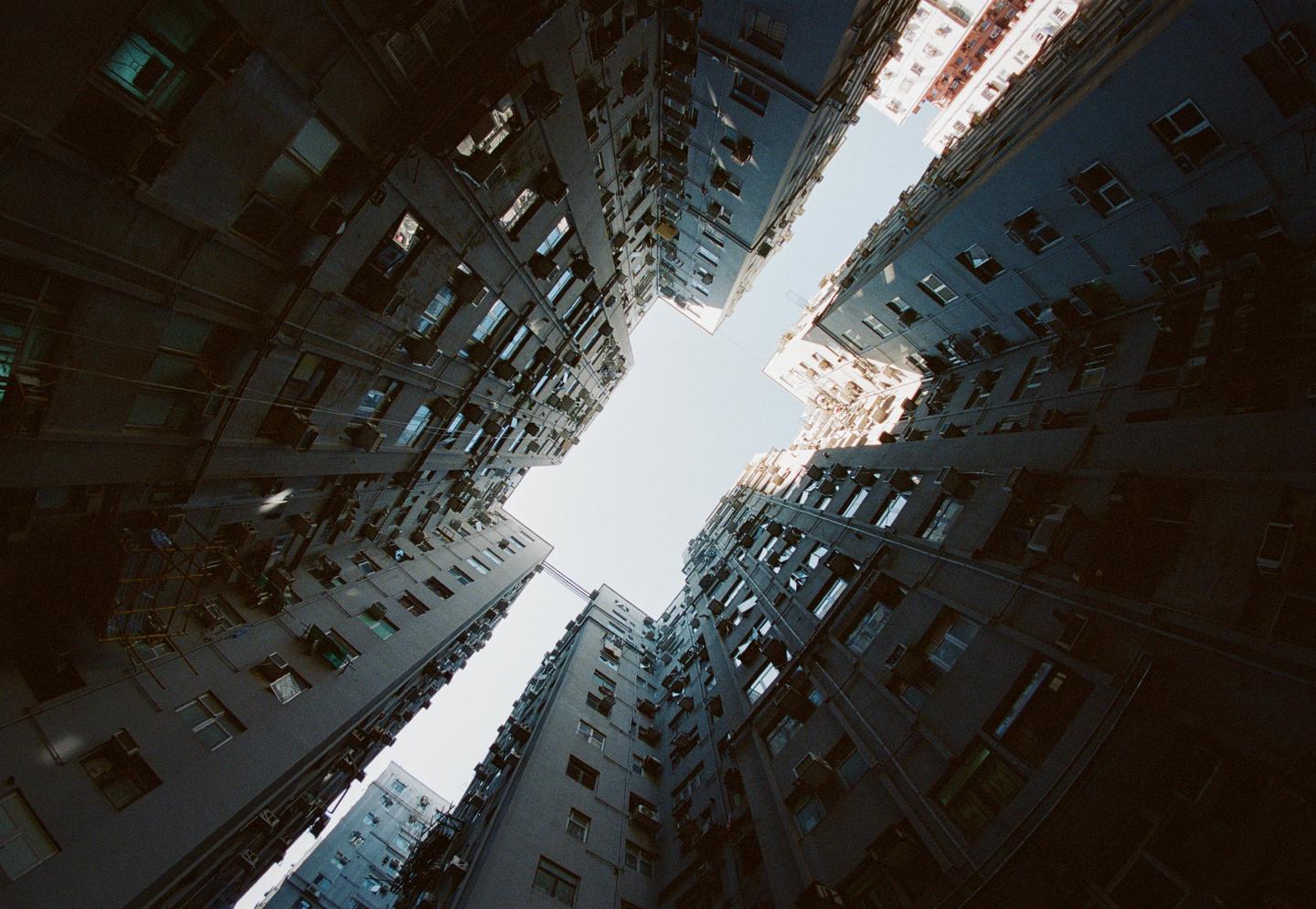 Looking up from a narrow courtyard, tall apartment buildings with many windows and air conditioning units rise towards a bright sky.