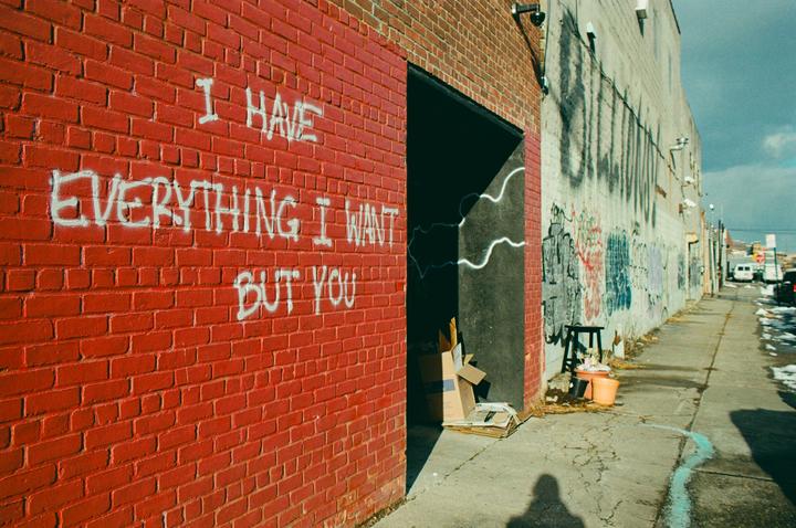 White graffiti on a red brick wall reads "I HAVE EVERYTHING I WANT BUT YOU" next to an alleyway with more graffiti and a shadow of a person.