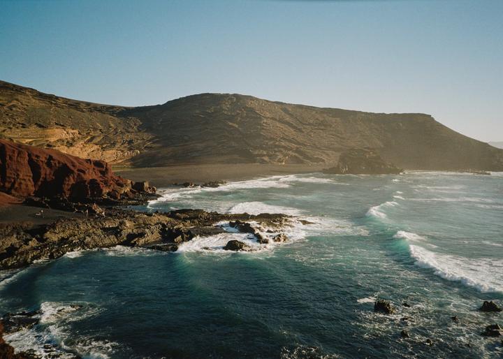 Waves crash against a rocky coastline with a dark sand beach and mountains in the background.
