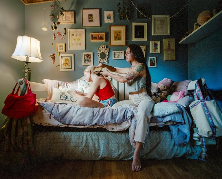 A woman combs a girl's hair while they sit on a bed in a room with many framed pictures on the wall.