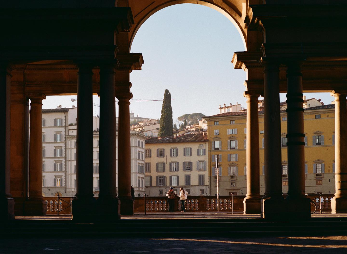 Two women stand on a balcony overlooking a city with buildings and a tall tree, framed by an archway and columns.
