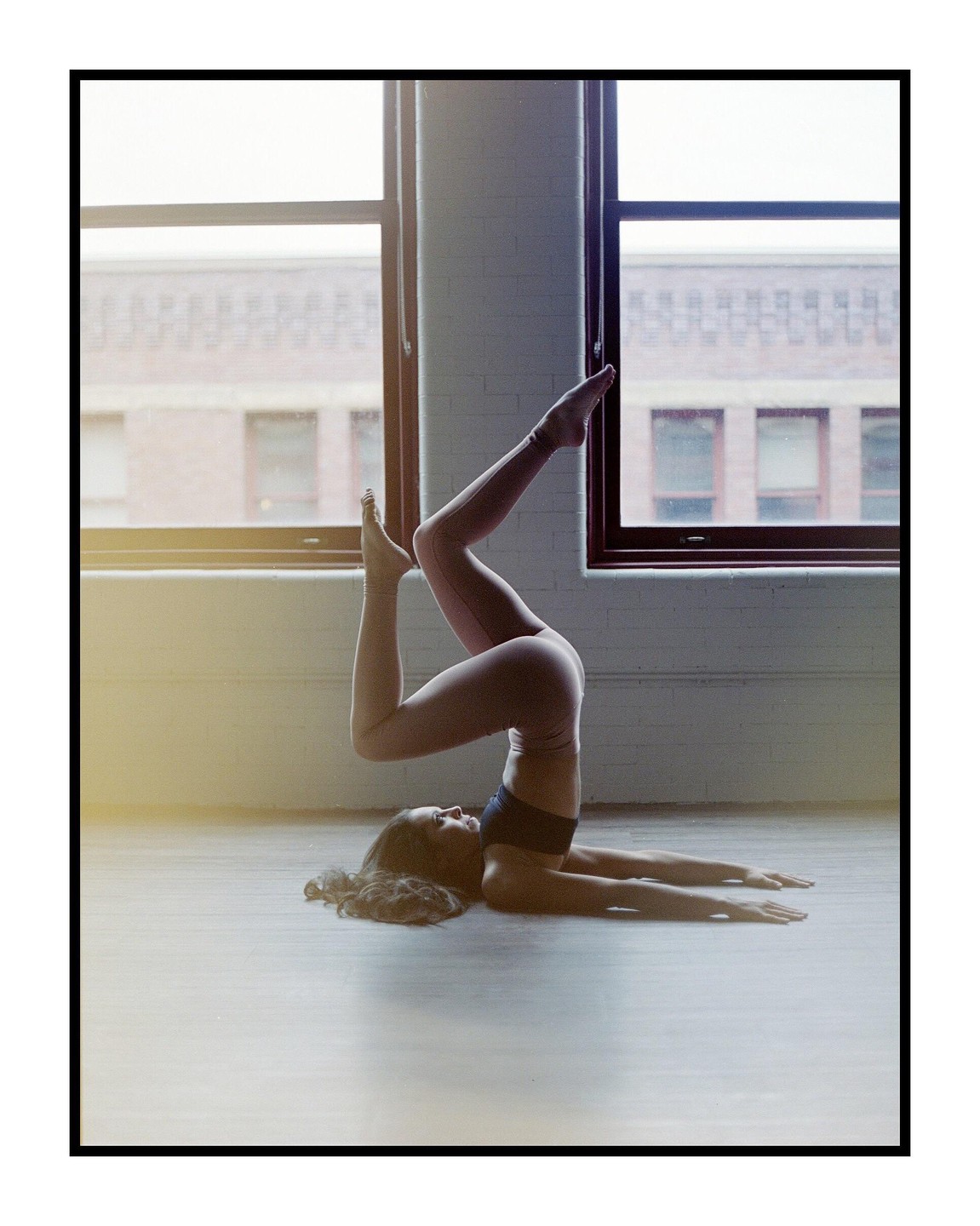 A woman in athletic wear performs a yoga pose on a wooden floor in a room with large windows.