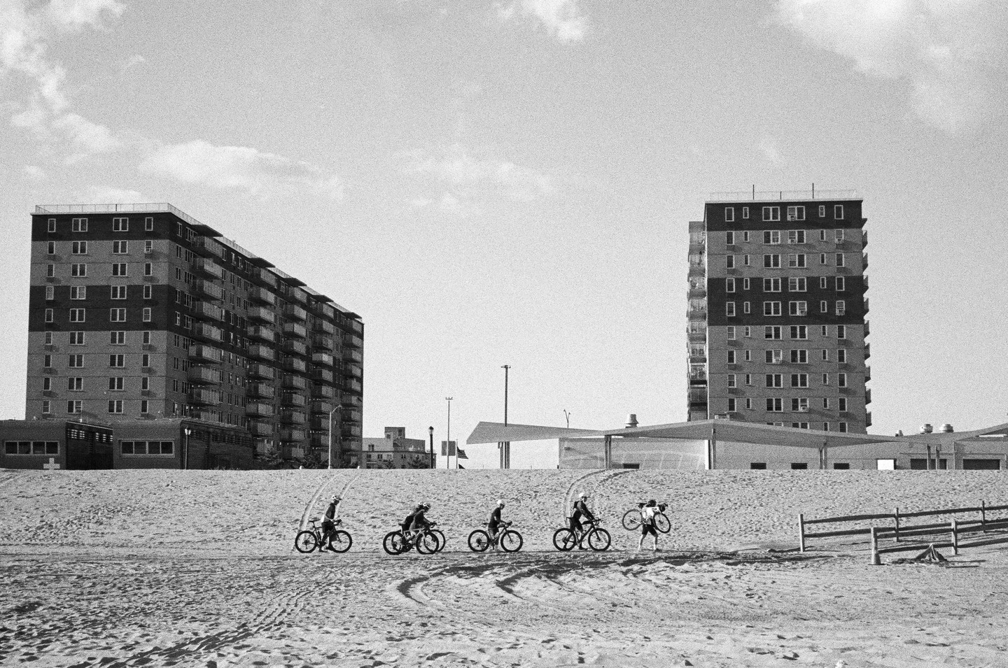 Four cyclists ride on a sandy beach with large buildings in the background, while one person carries a bicycle over their shoulder.