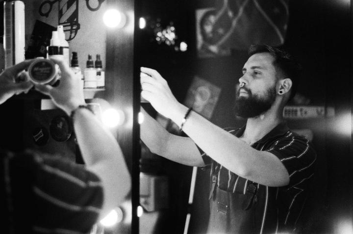 A man with a beard and nose ring is reflected in a mirror, reaching for something on a shelf.