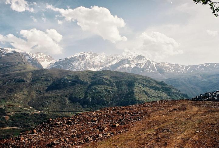 A rocky, reddish-brown foreground leads to green hills and snow-capped mountains under a cloudy sky.