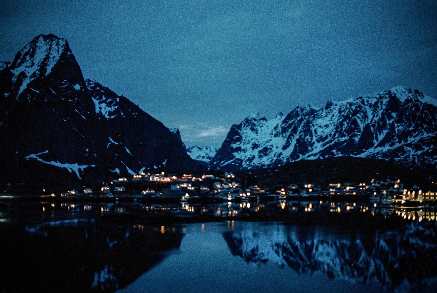 A small town with illuminated buildings sits at the base of snow-capped mountains, reflected in the still water of a lake at night.