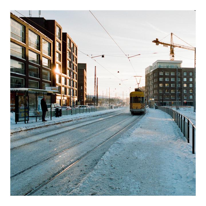 A man stands at a snowy tram stop with a tram approaching in the distance, surrounded by buildings and overhead wires.