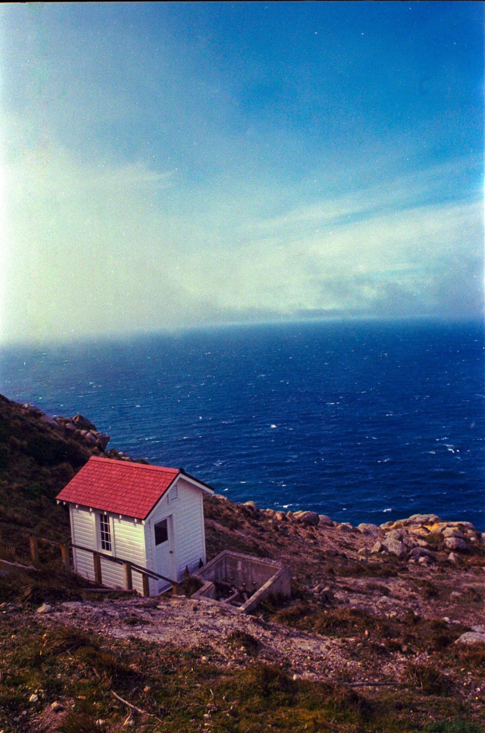 A small white house with a red roof sits on a grassy cliff overlooking a vast blue ocean under a hazy sky.