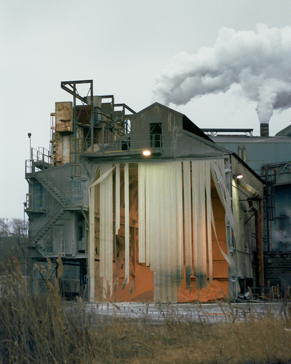 A large industrial building with smoke billowing from a smokestack stands behind tall dry grass.