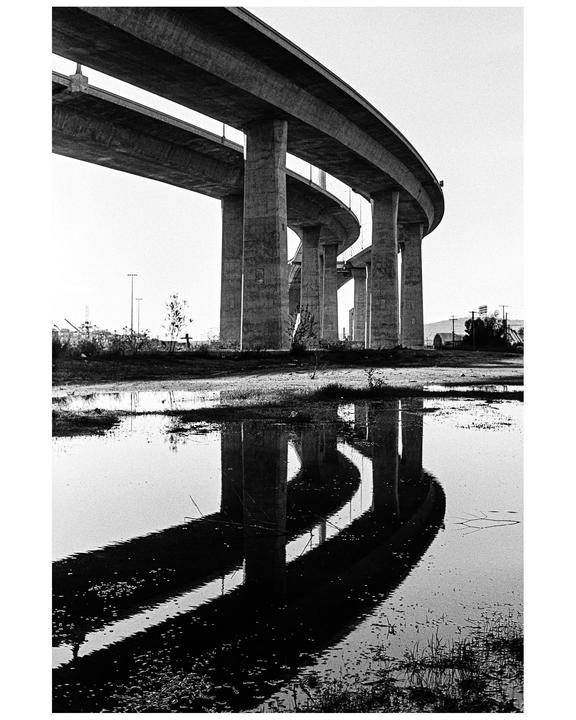 A concrete overpass with multiple levels is reflected in a large puddle on the ground.