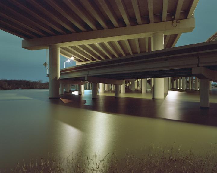 A highway overpass with many support pillars stands over a body of water at night.