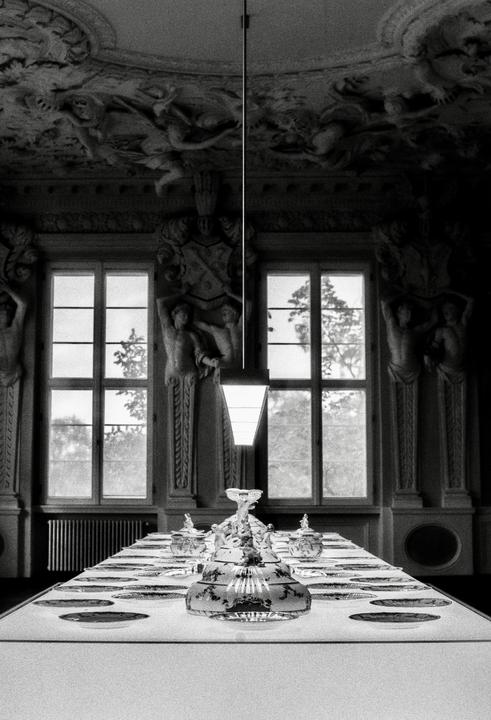A long dining table set with plates and glassware sits in a grand room with ornate ceilings and large windows.