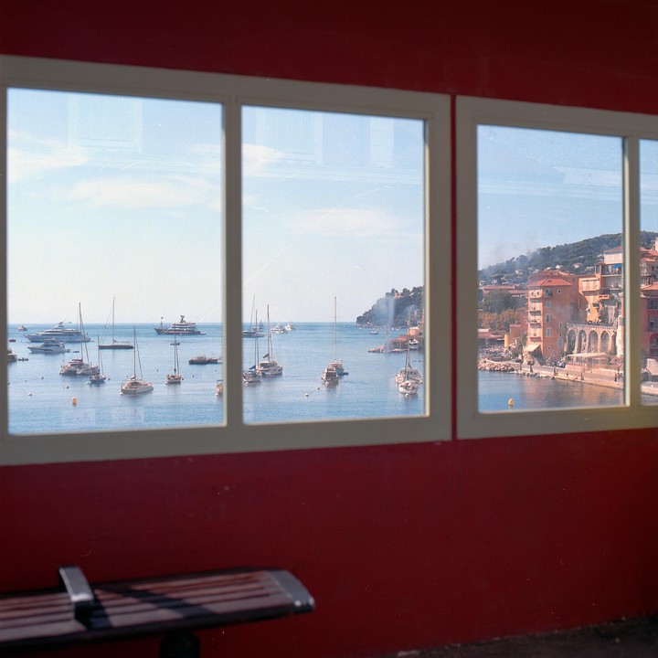 A view of a harbor filled with boats and a town built into a hillside is seen through a window with a red wall below.