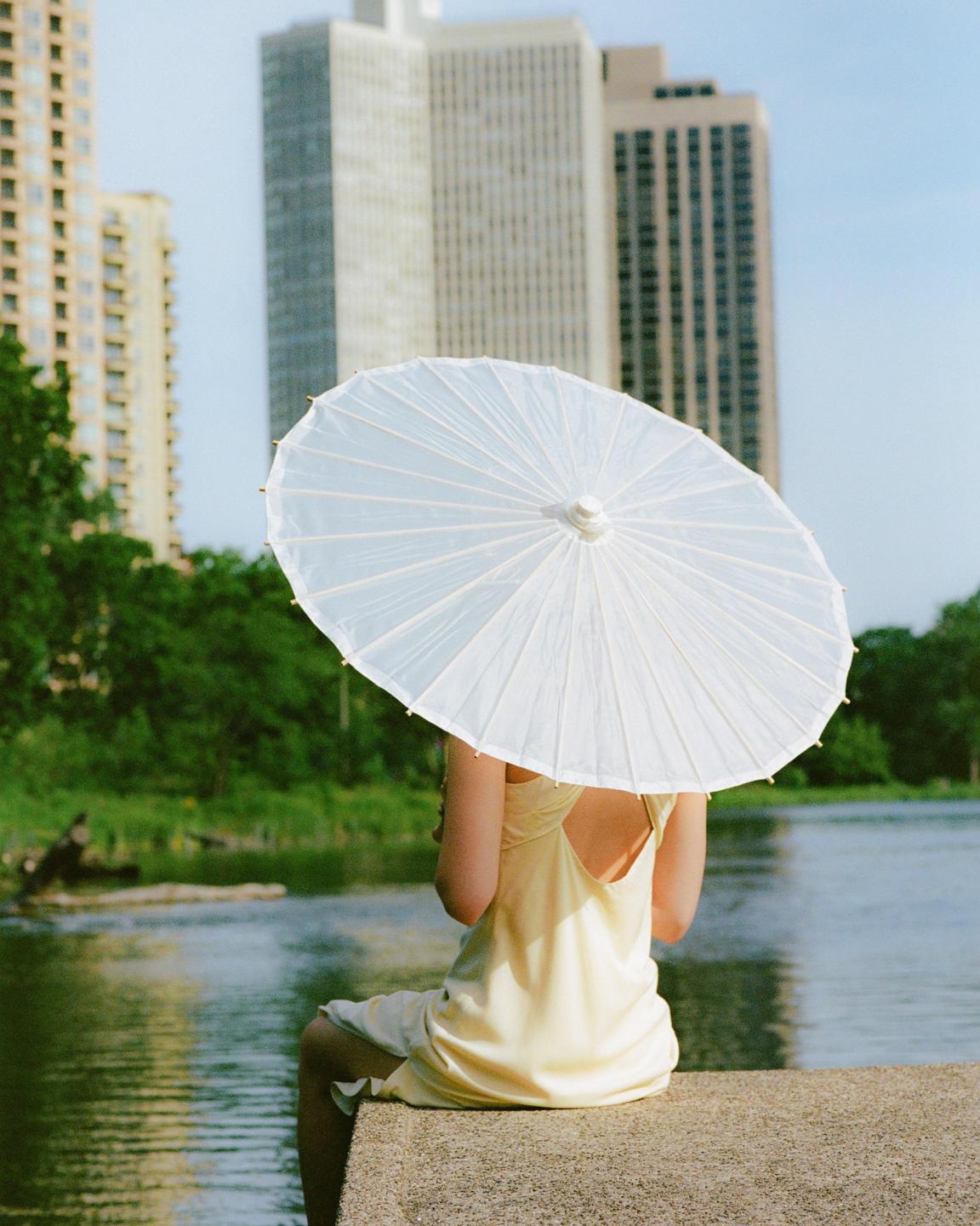 A woman in a yellow dress sits by a body of water, holding a white parasol, with tall buildings in the background.