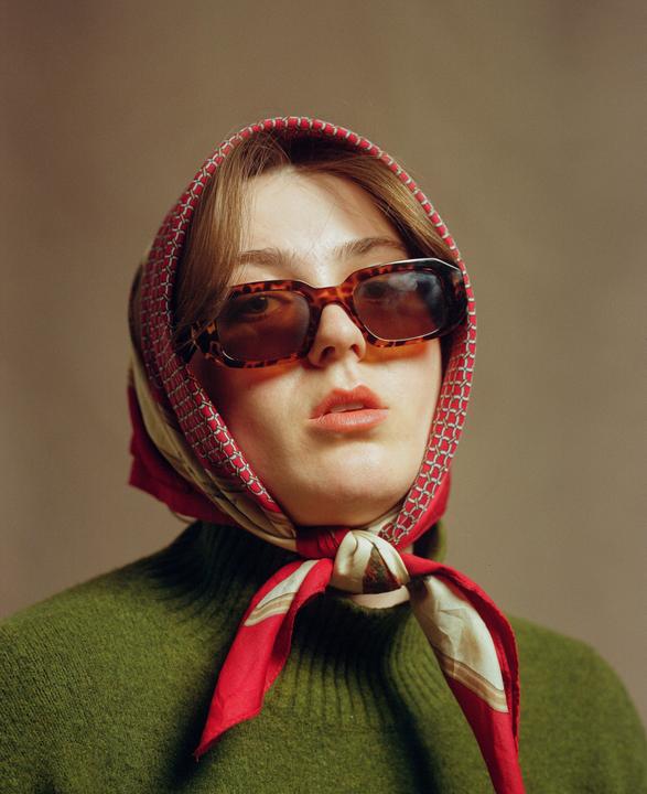 A woman wearing a red and white headscarf, sunglasses, and a green sweater looks directly at the viewer.