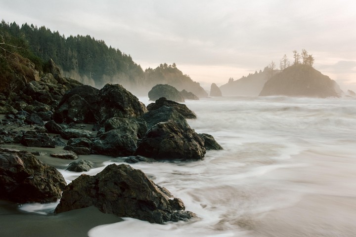 Waves crash on a rocky beach with forested cliffs and sea stacks in the background.