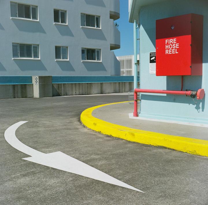 A white arrow is painted on an asphalt surface next to a bright yellow curb and a light blue building with a red fire hose reel.
