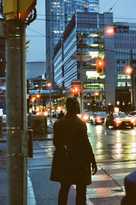 A man stands at a crosswalk in a city at dusk, with cars and buildings visible in the background.