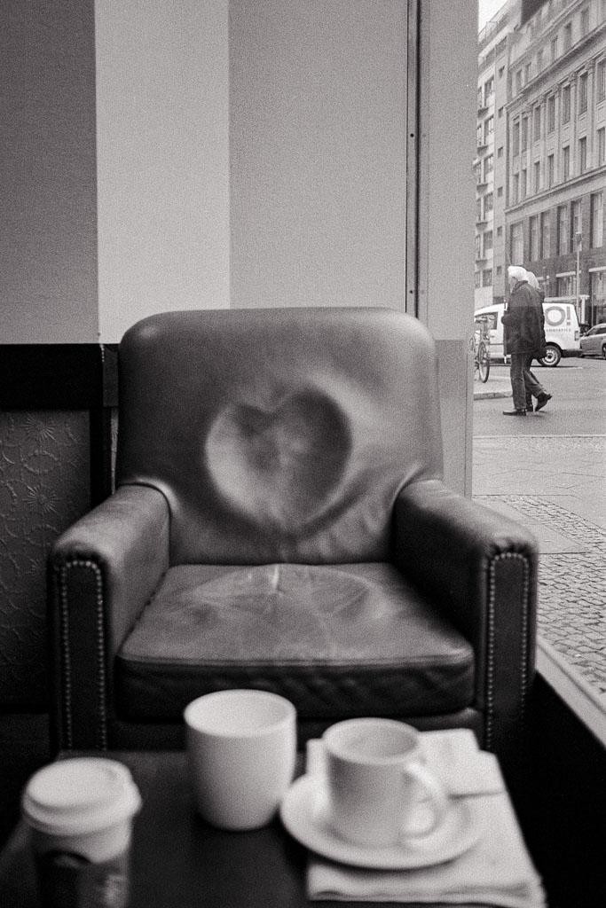 A leather armchair with a heart-shaped indentation on its back is next to a table with three cups, with a street scene visible through a window in the background.