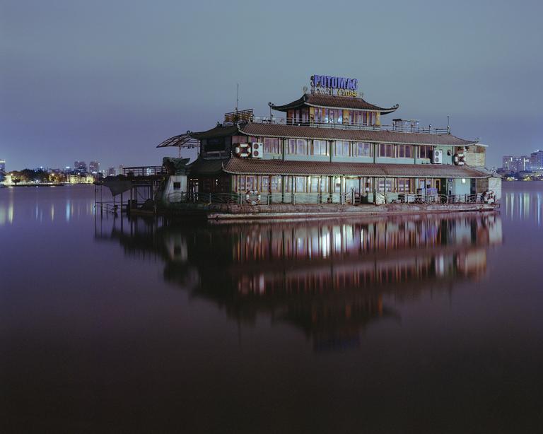 A large boat with a pagoda-style roof is illuminated at night, reflecting on the still water.