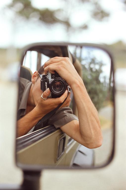 A man is reflected in a side mirror, holding a camera to his eye.