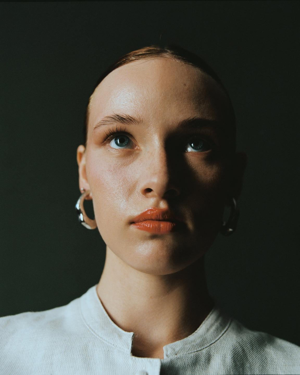A woman with blue eyes and orange lipstick looks up, wearing a light-colored shirt and hoop earrings.
