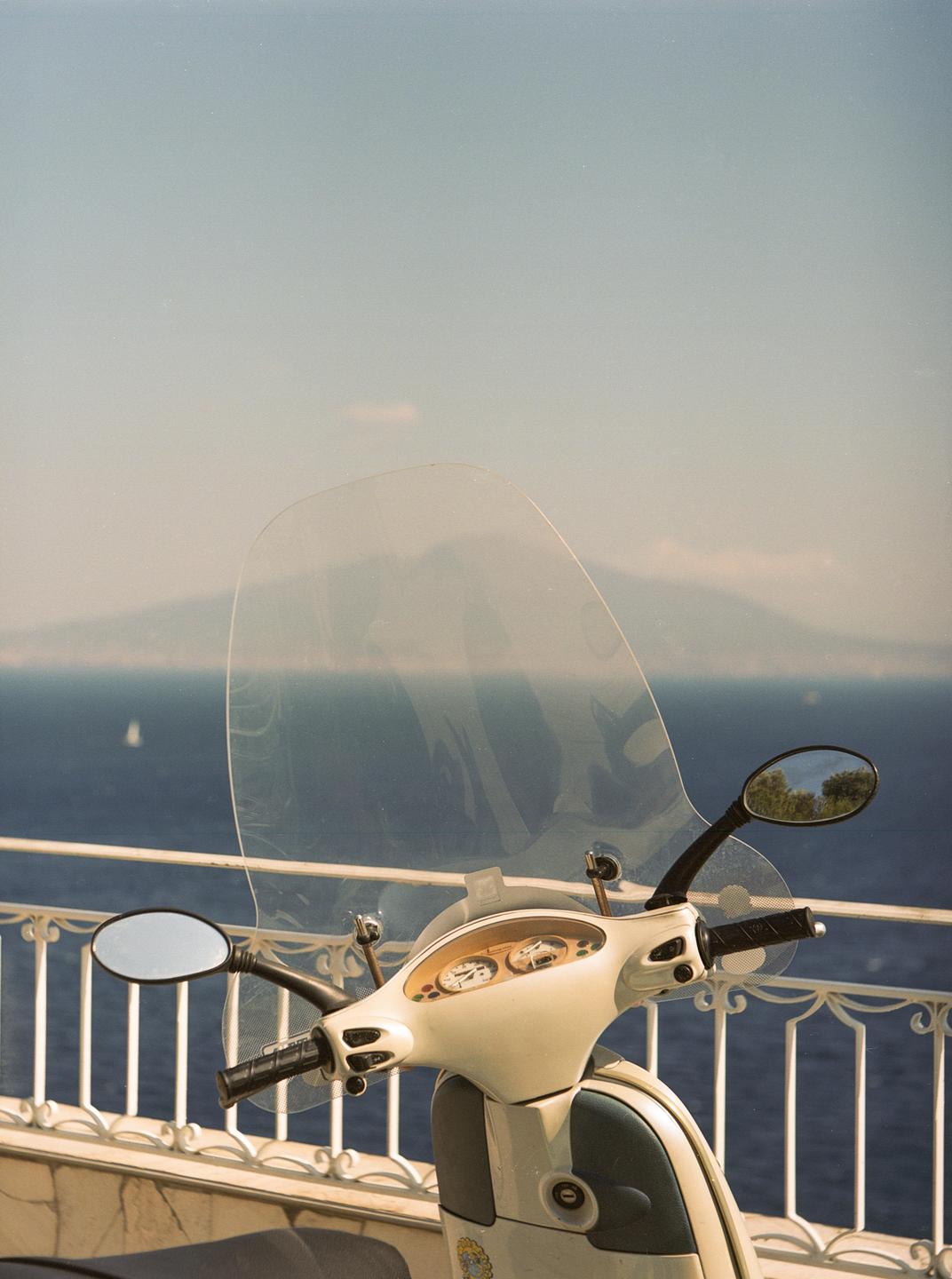 A scooter is parked on a balcony overlooking the sea and a distant mountain.
