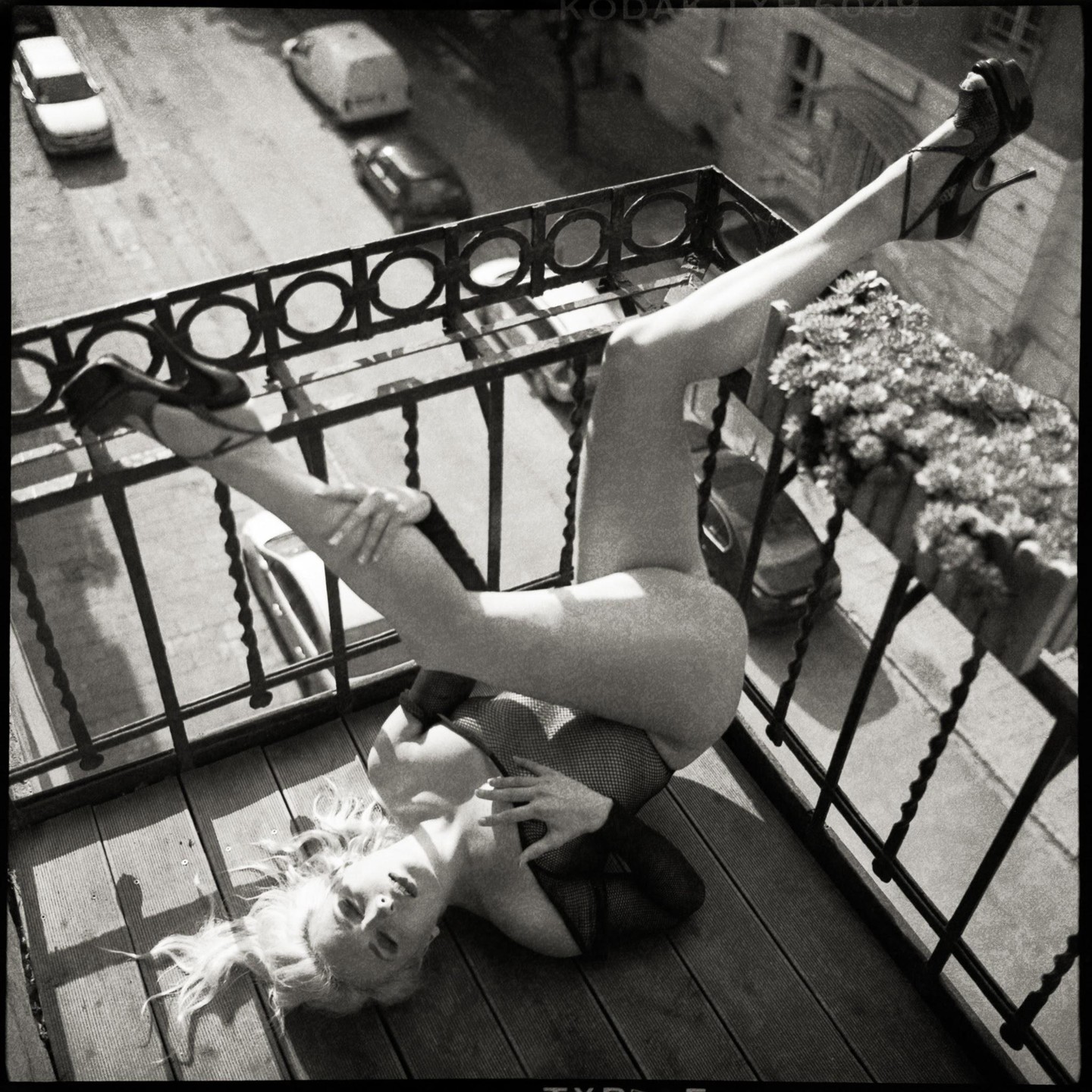 A woman in fishnet lingerie and high heels is lying on a balcony with her legs up on the railing.