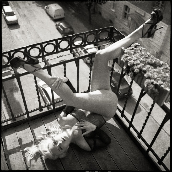 A woman in fishnet lingerie and high heels is lying on a balcony with her legs up on the railing.