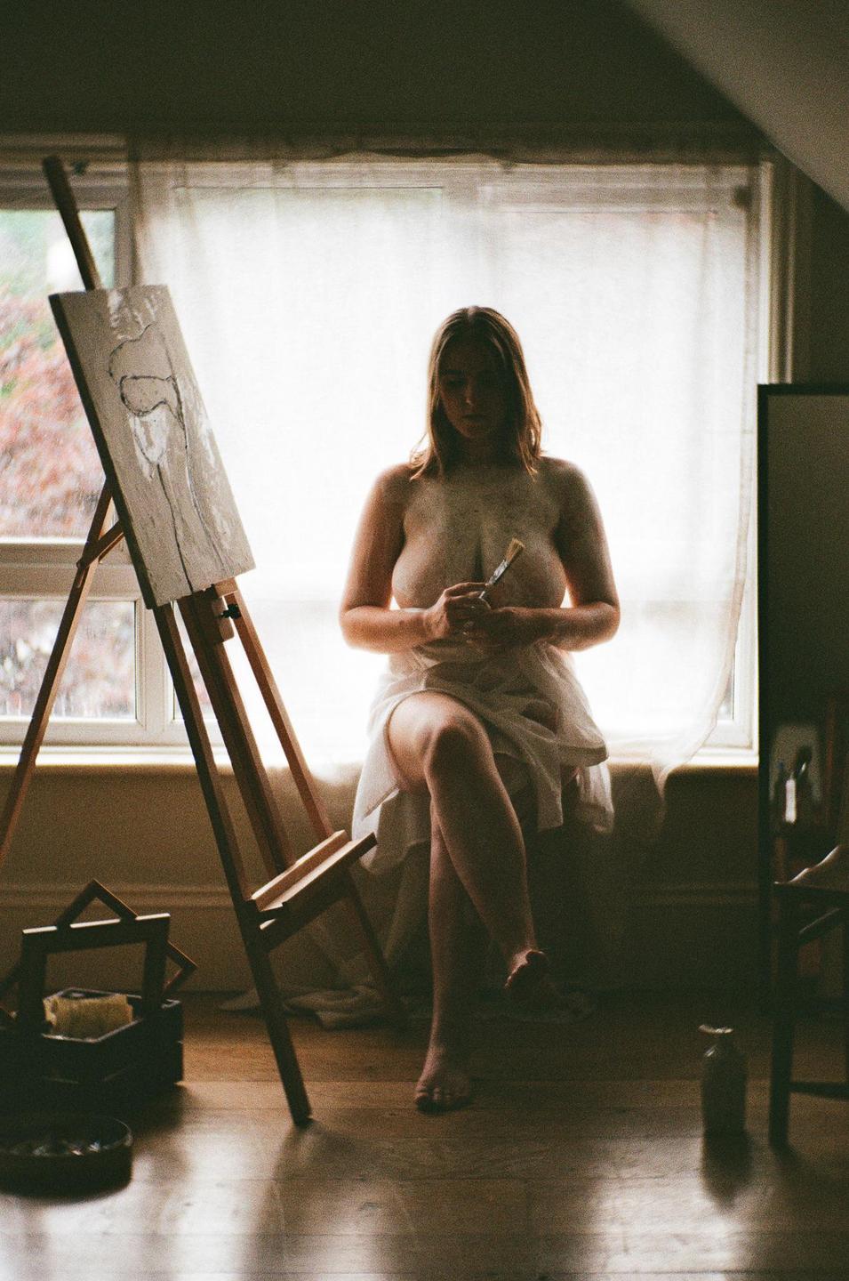 A woman sits on a window sill, holding a paintbrush, next to an easel with a drawing.
