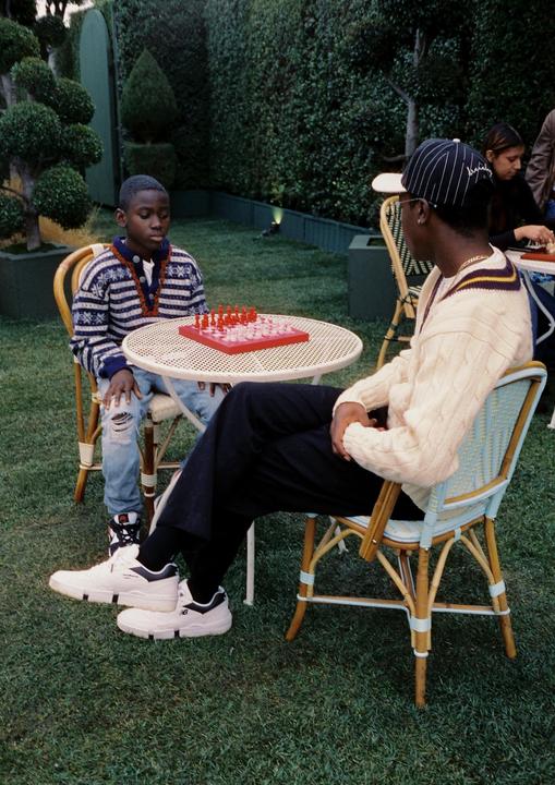 A boy and a man play chess at a small table on a grassy lawn surrounded by hedges.