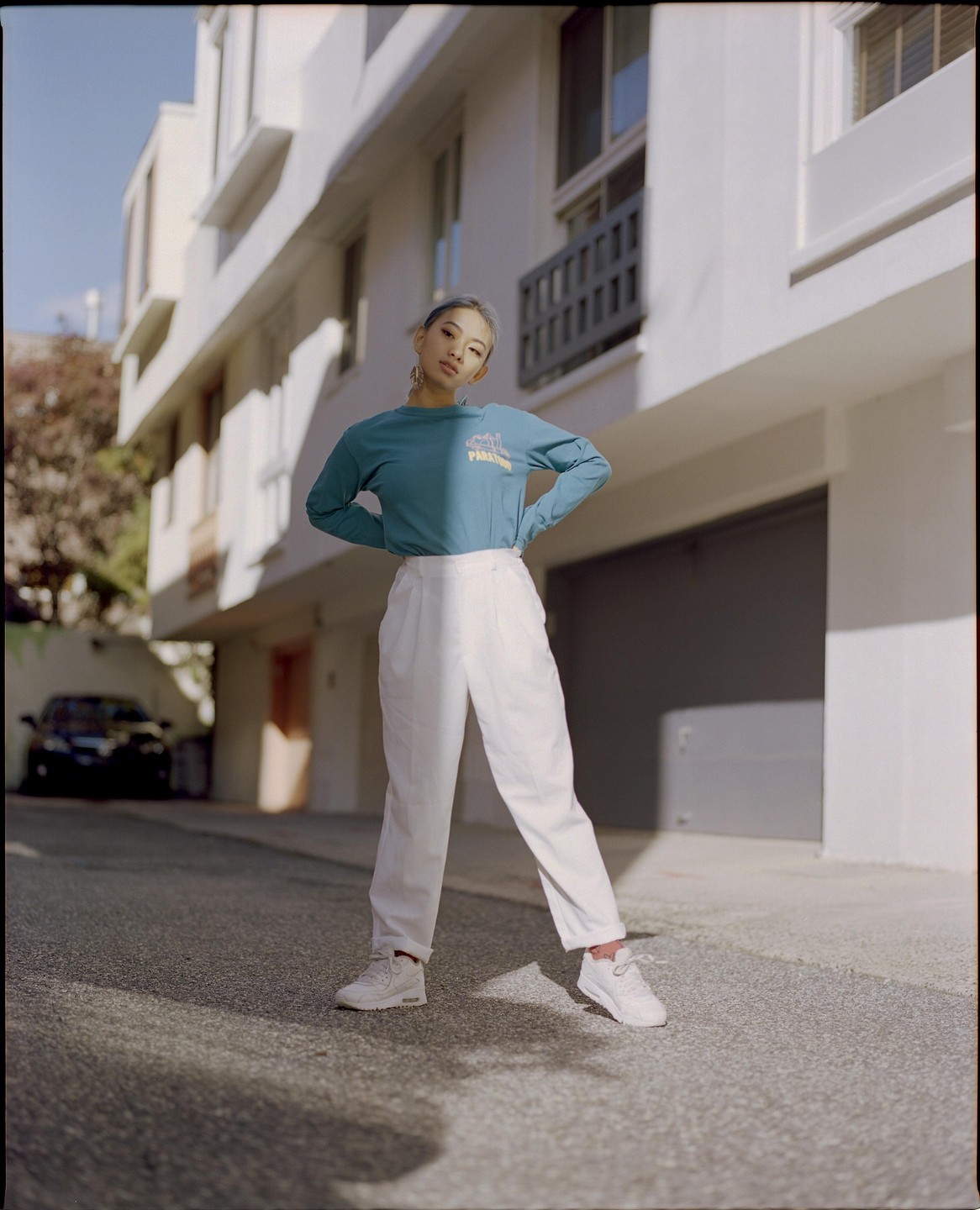 A woman with blue hair stands on a paved surface in front of a white building with her hands on her hips.