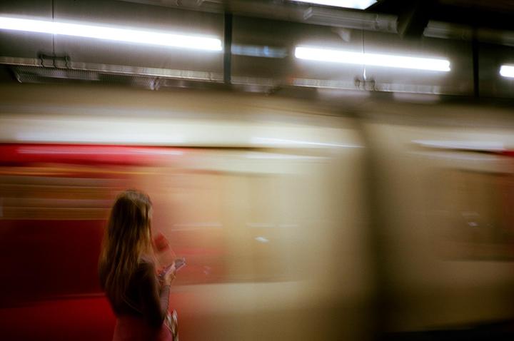 A woman stands on a subway platform with a blurred train passing by.