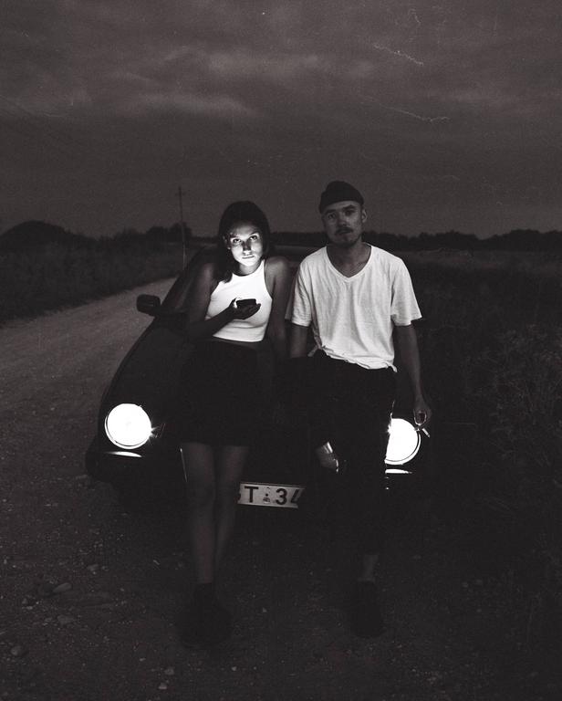 A couple stands in front of a car with its headlights on, with the woman looking at her phone.