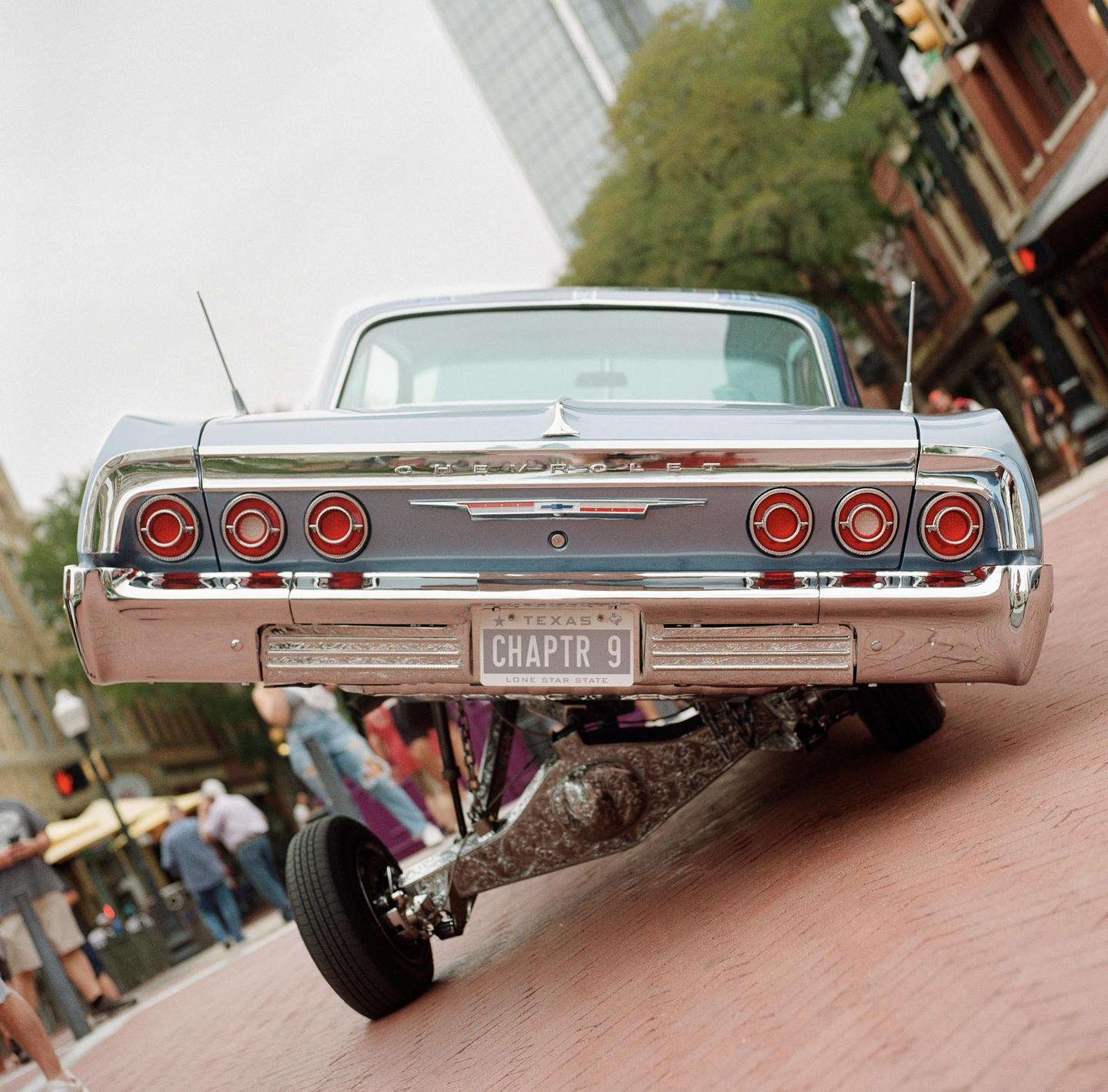 A blue lowrider car with a Texas license plate is parked on a brick street.