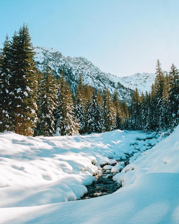 A stream flows through a snow-covered valley surrounded by pine trees and mountains.