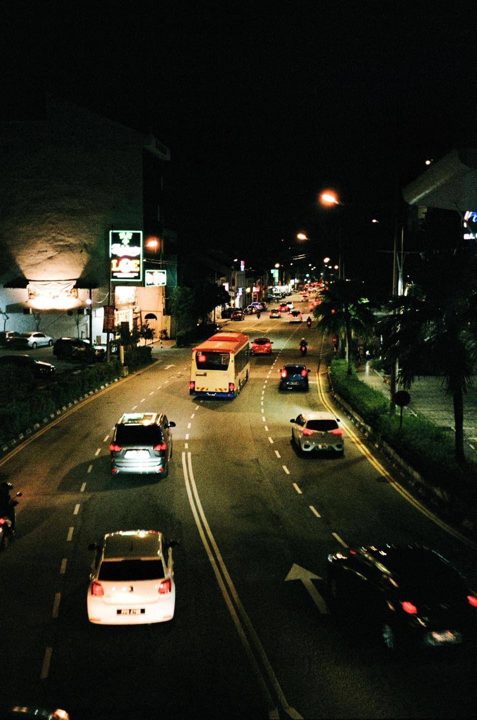 A street at night is filled with cars, a bus, and motorcycles, with buildings and palm trees lining the road.
