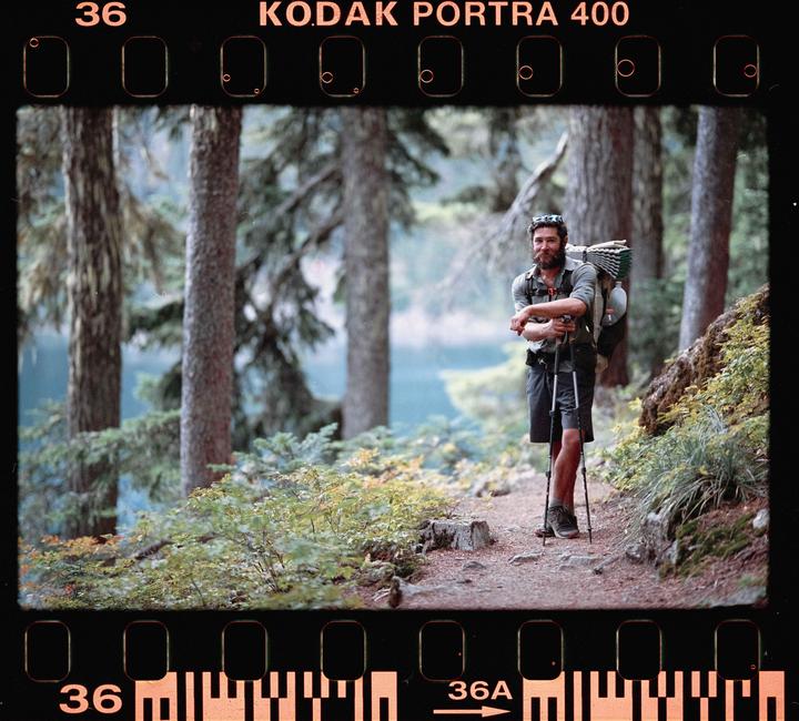A man with a beard and backpack stands on a trail in a forest next to a body of water.